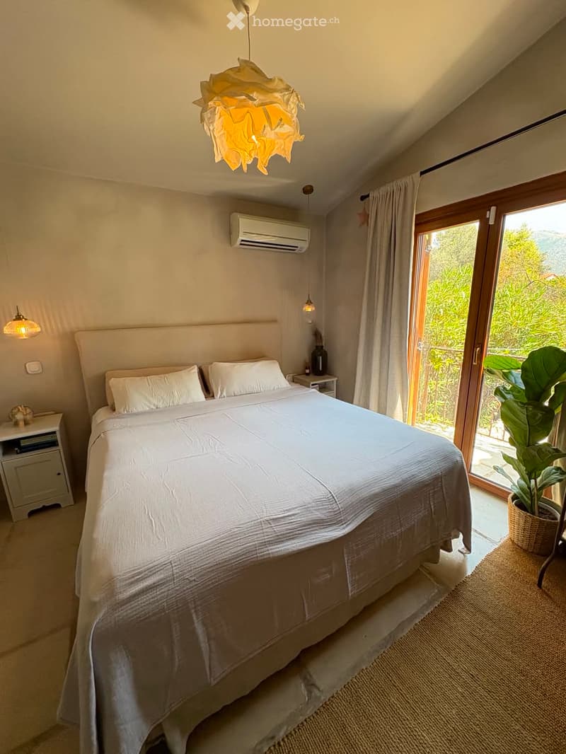 Modern bedroom with a large bed, nightstands, a hanging lamp, and a view through large windows.