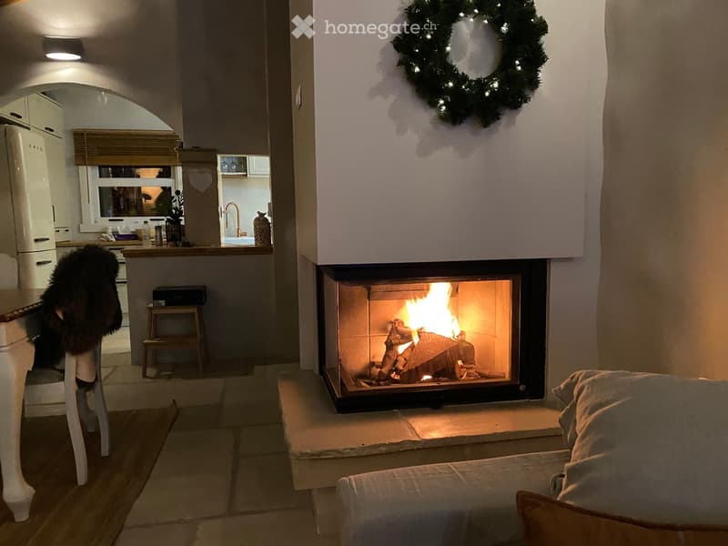 Cozy living room with a lit fireplace, a sofa, a small stool, and a kitchen area visible in the background.
