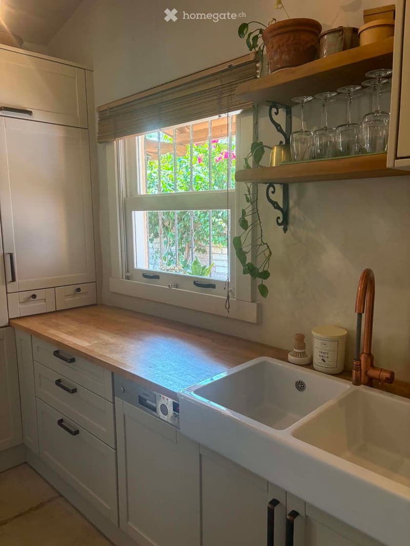 A modern kitchen with a double sink, wooden countertop, white cabinets, and a window overlooking a garden. Shelves hold glassware and a potted plant.