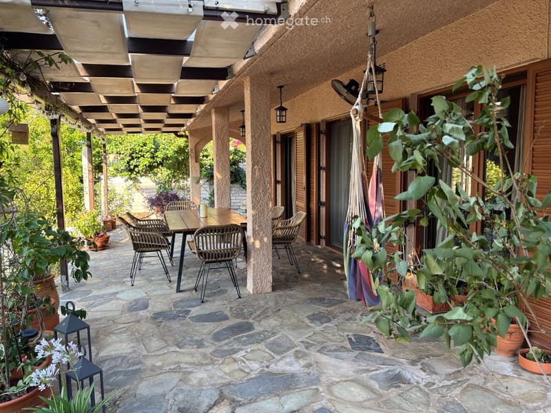 A covered outdoor patio with stone flooring, wooden furniture including a table and chairs, potted plants, and hanging lanterns.