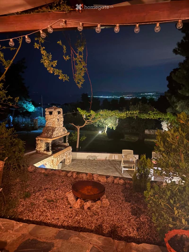 Outdoor patio with a stone fire pit, a stone barbecue, a chair, string lights, and a scenic night view of the city in the background.