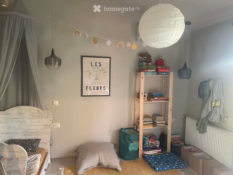 A cozy children's bedroom with a wooden bed, a shelf filled with books and toys, a large round paper lantern, and a framed picture on the wall. The room features a curtained bed area, a small rug, and a storage box on the floor.