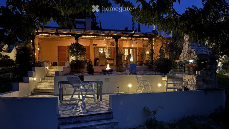 Evening view of a well-lit outdoor patio area with a stone fire pit, a table with chairs, and steps leading up to a covered terrace with additional seating and lighting.