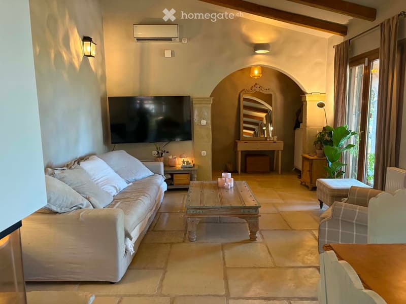 Living room with a sofa, coffee table, chair, mirror, and television. The room features arched doorways and warm lighting.