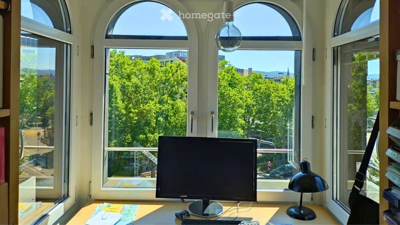 Bright room with large arched windows offering a view of greenery and cityscape. A wooden desk holds a computer monitor, keyboard, mouse, and a black desk lamp. A bookshelf with books is visible on the right side.