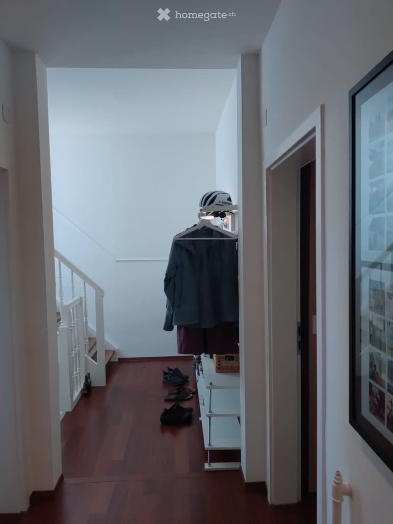 A narrow hallway with wooden flooring, white walls, and a staircase leading upwards. A coat and helmet are hanging on a rack, and several pairs of shoes are placed on the floor near the staircase.