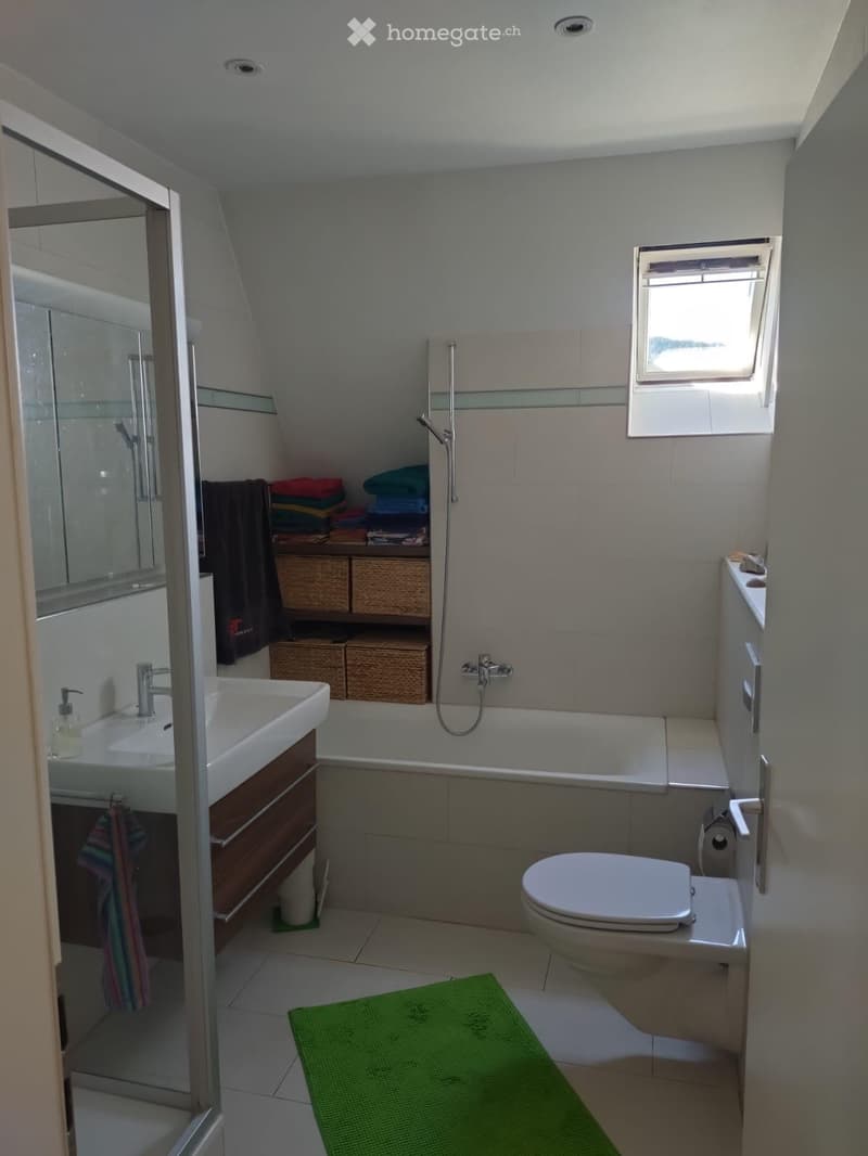 Modern bathroom with a white sink, toilet, bathtub, and shower area. Features a large mirror, towel rack, and a green bath mat on the floor.