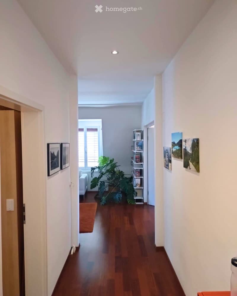A well-lit hallway with wooden flooring, white walls, and several framed pictures. A small plant and a shelf are visible in the background, along with a partially visible living room area.