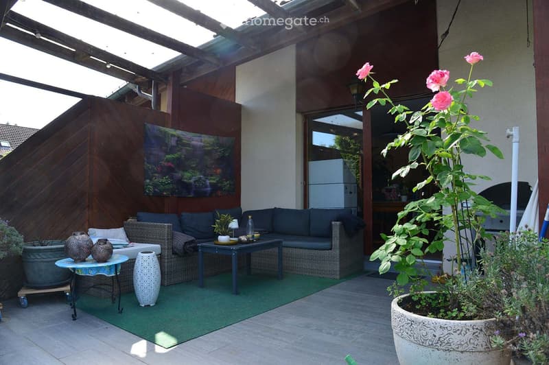 Outdoor patio with a green artificial turf, wicker sofa set, small tables, decorative vases, and potted plants including a large rose bush.