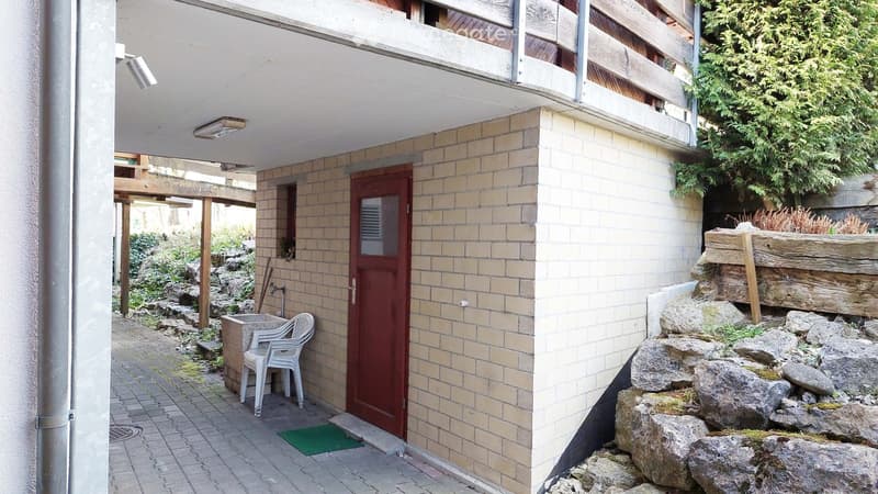 Small outdoor area with a brick wall, a red door, a white plastic chair, and a small garden with rocks and greenery.