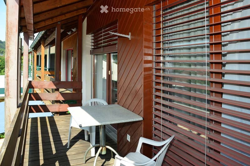 Balcony with wooden decking, white plastic table and chairs, wooden shutters, and a view of the surrounding area.