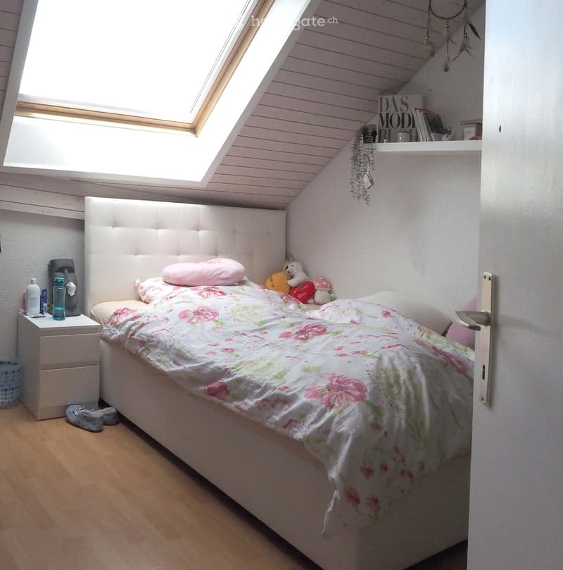 Cozy bedroom with a white upholstered bed, floral duvet, nightstand, and decorative items including toys and a dreamcatcher.