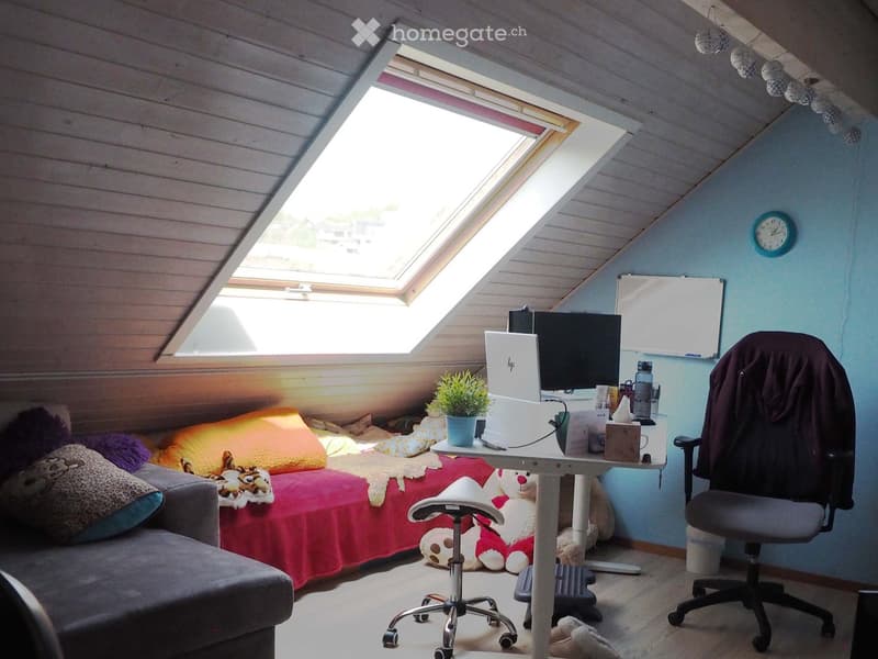 Cozy attic room with slanted wooden ceiling, skylight window, bed with colorful blankets, desk with laptop and chair, and decorative items.