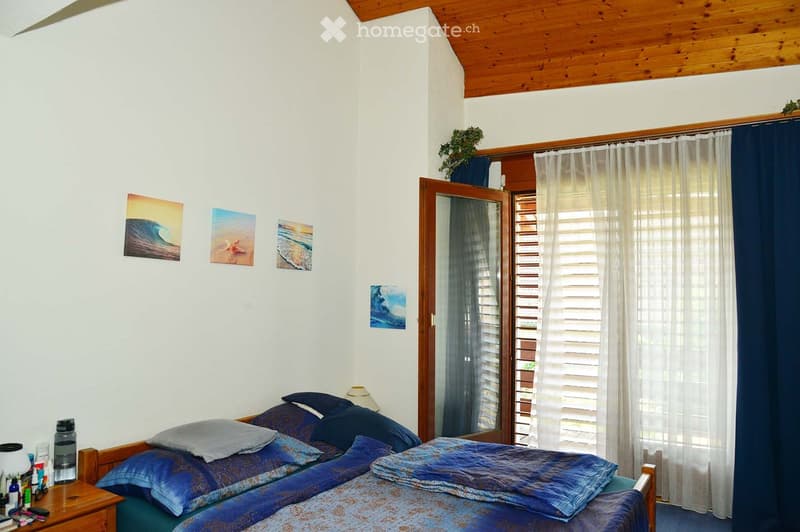 Cozy bedroom with a double bed, nightstand, lamp, mirror, and artwork on the walls. The room features a wooden ceiling and a large window with curtains.