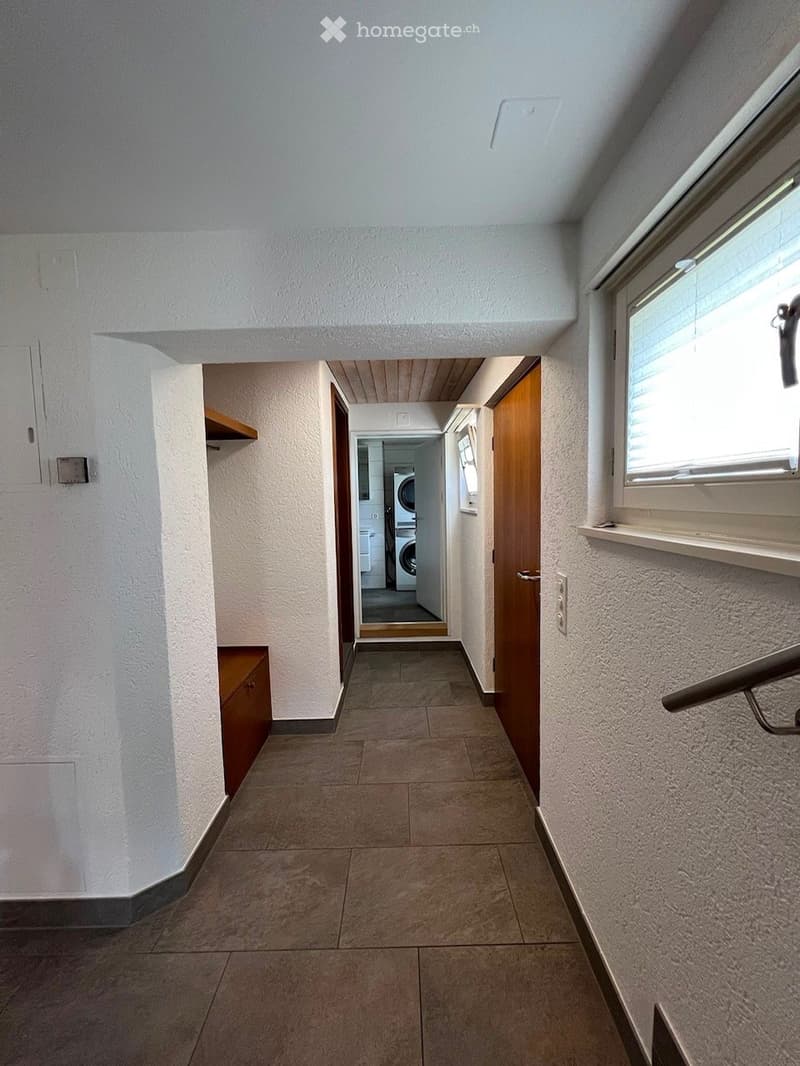 A clean, well-lit hallway with tiled flooring, white walls, and wooden doors leading to other rooms. A small wooden cabinet is visible on the left side, and a window with blinds is on the right.