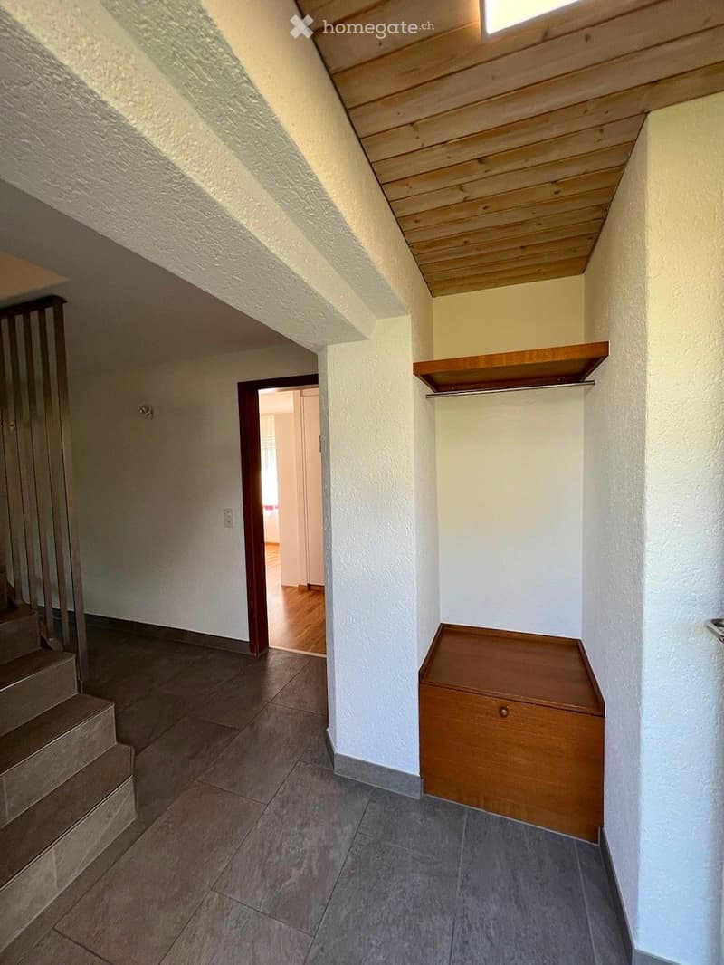 Empty hallway with tiled floor, white walls, wooden shelf, and a small wooden bench. A staircase leads upwards, and an open doorway reveals another room with wooden flooring.