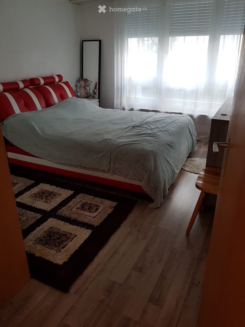 Bedroom with a red and white upholstered bed, wooden floor, patterned rug, nightstand, and mirror.