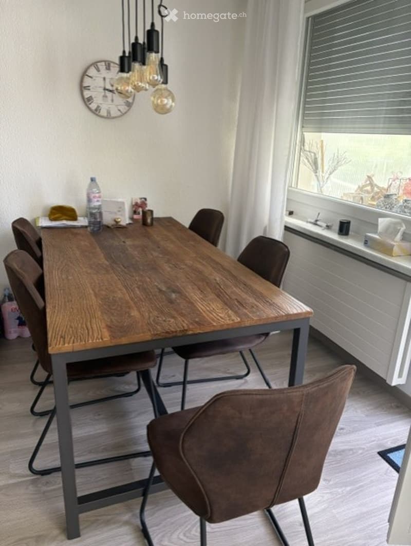 Dining room with a wooden dining table, six chairs, a clock on the wall, and a bottle on the table.
