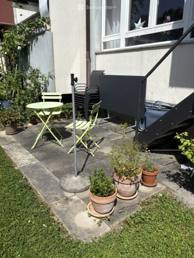 Small outdoor patio with a round table, four green chairs, several potted plants, and a black barbecue grill.