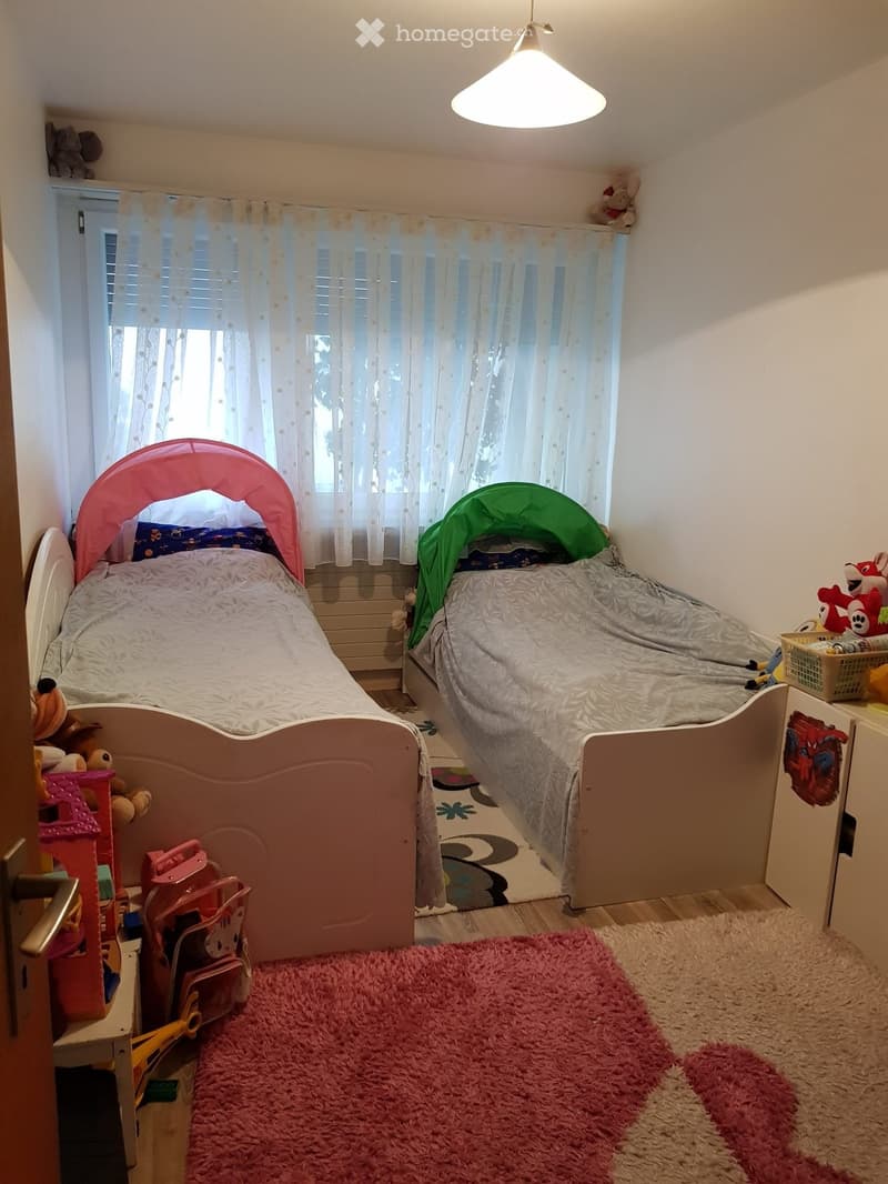 Cozy children's bedroom with two single beds, one with a pink canopy and the other with a green cover, surrounded by toys and a soft pink rug.