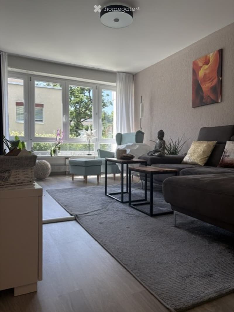 Modern living room with large windows, grey sofa, coffee table, side table, chair, floor lamp, and decorative items including a plant and a statue.