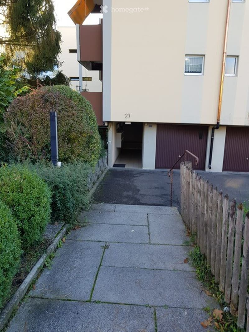 Side view of a multi-story residential building with a garage entrance, concrete pathway, and greenery on either side.