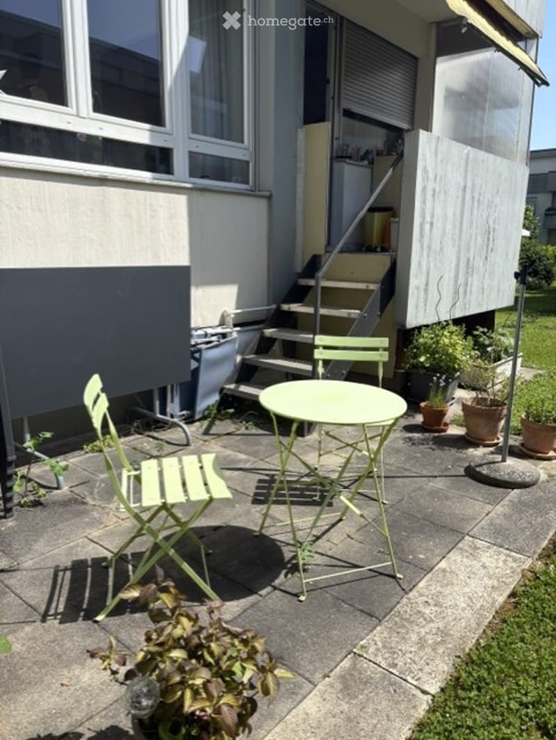 A small outdoor patio area with a round table, two folding chairs, and several potted plants. The patio is adjacent to a building with a staircase leading up to an upper level.