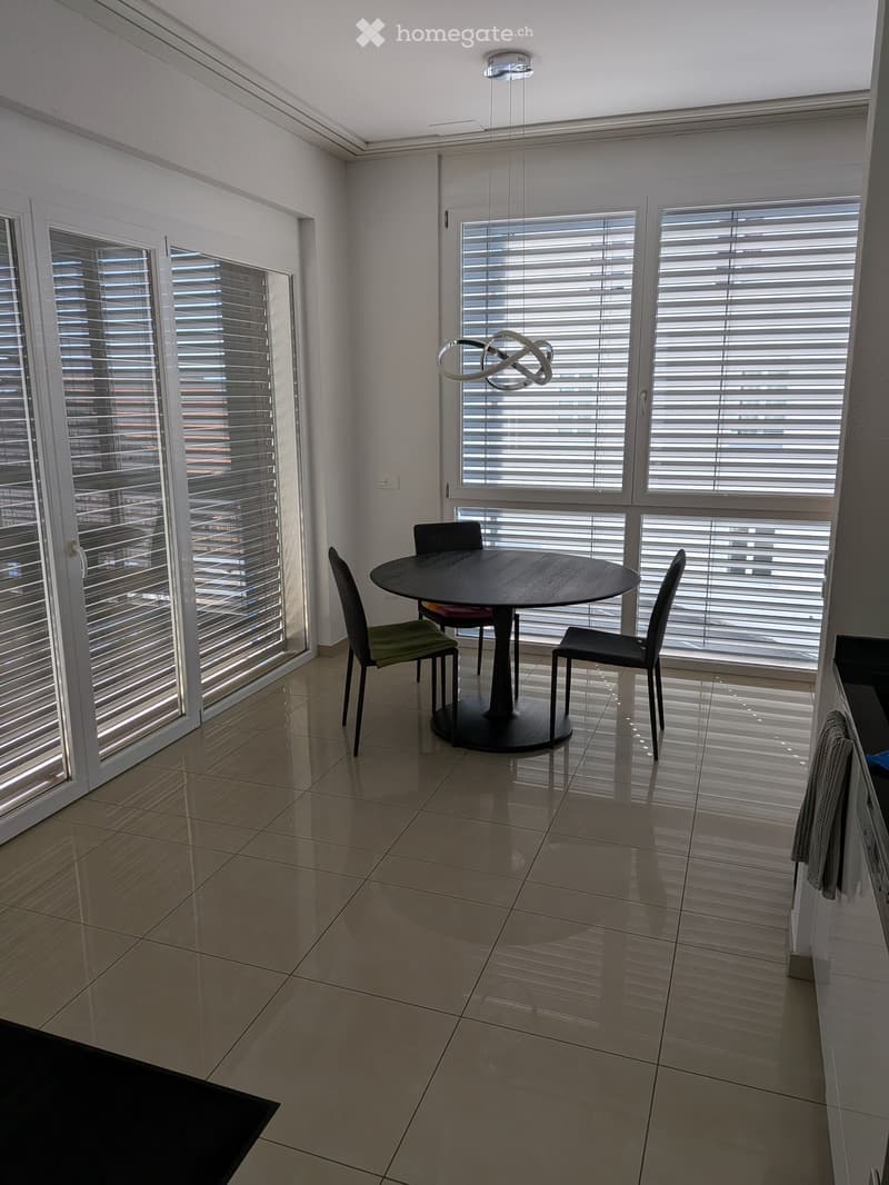 Modern dining area with large windows, a round wooden table, four chairs, and a contemporary light fixture.
