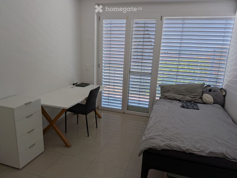 A modern, compact bedroom with a single bed, a desk with a chair, and a drawer unit. The room features large windows with blinds and a tiled floor.