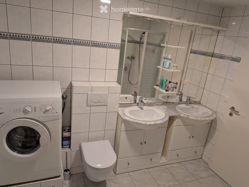 Modern bathroom with white tiles, a double sink vanity, a toilet, a washing machine, and a glass-enclosed shower area.