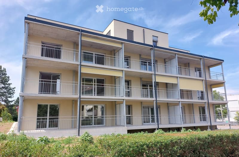 Modern multi-story residential building with beige and white facade, multiple balconies with metal railings, large windows, and a flat roof.