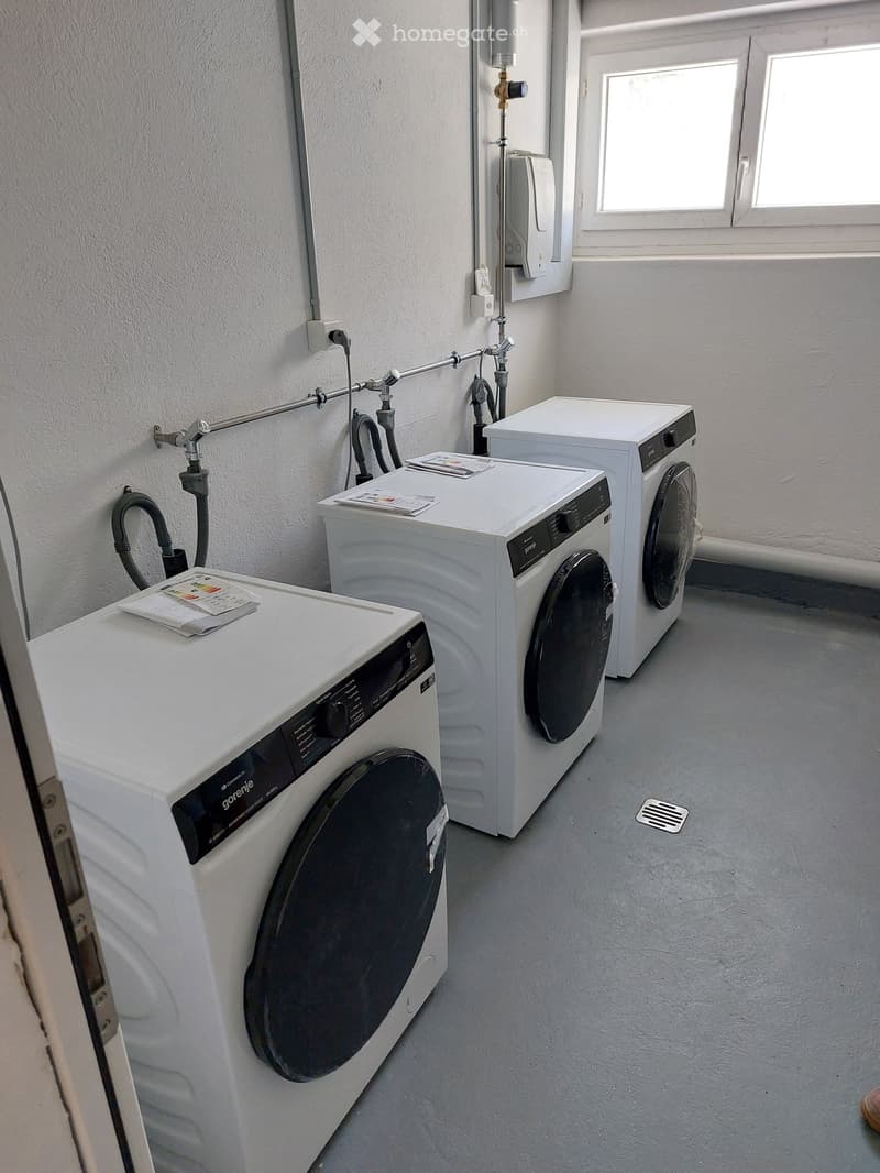 A small, well-lit room with three white washing machines and a dryer, featuring a window and a grey floor.