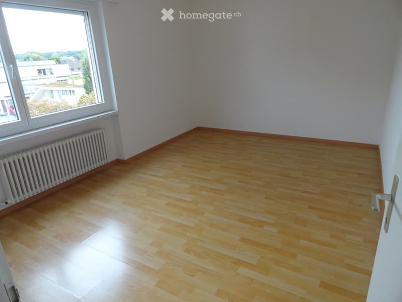 Empty room with wooden flooring, white walls, and a large window offering a view of the outside. A radiator is visible along the wall beneath the window.