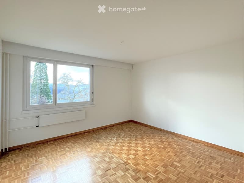 Empty room with wooden parquet flooring, white walls, and a large window with a view of trees.