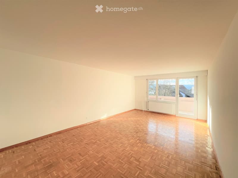 Empty room with wooden parquet flooring, white walls, and large windows with a sliding door leading to a balcony.