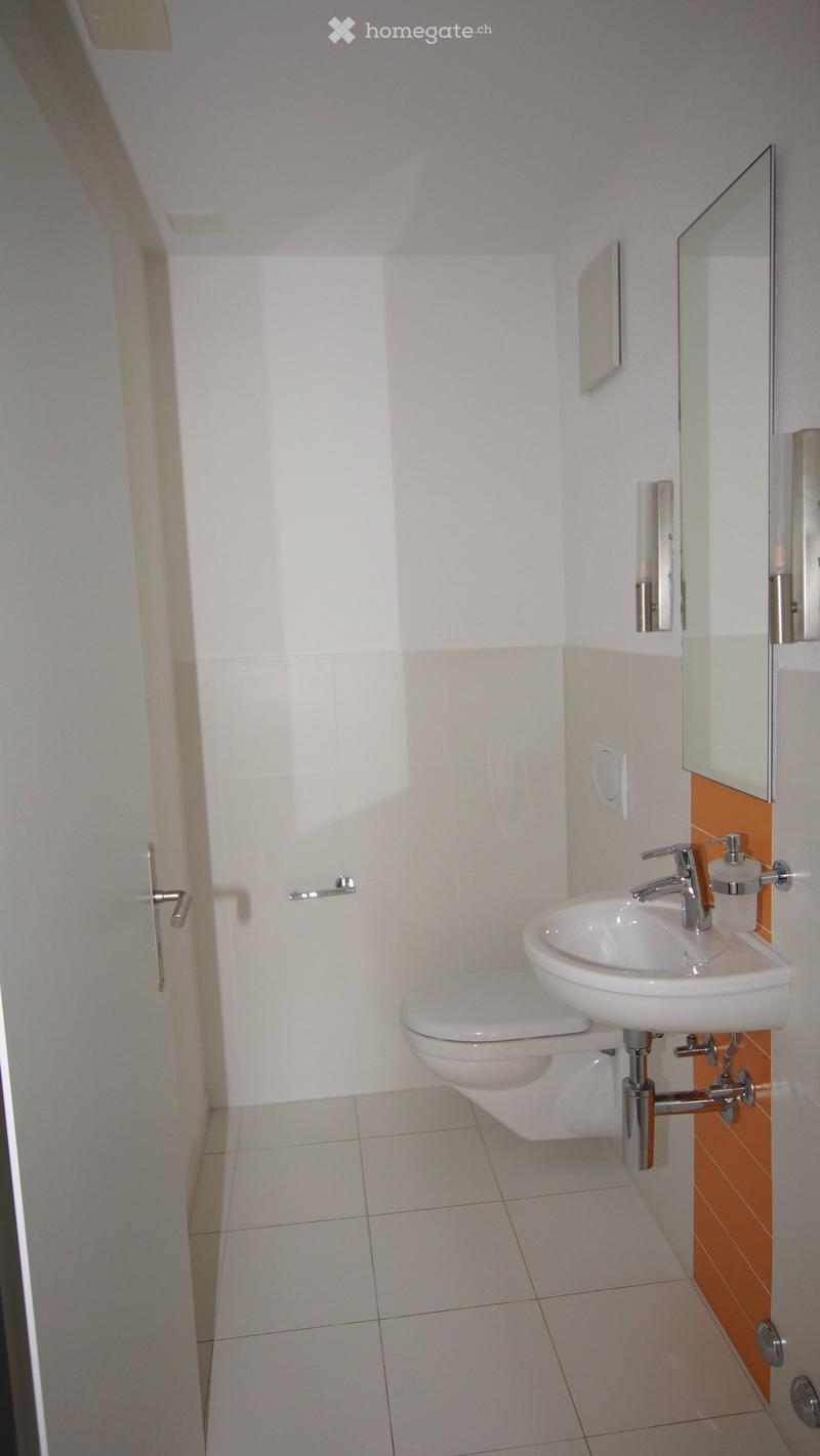 Modern bathroom with white tiled walls, a toilet, a sink with a chrome faucet, a mirror, and a soap dispenser. The floor is tiled in light gray, and the walls feature a contrasting orange accent.