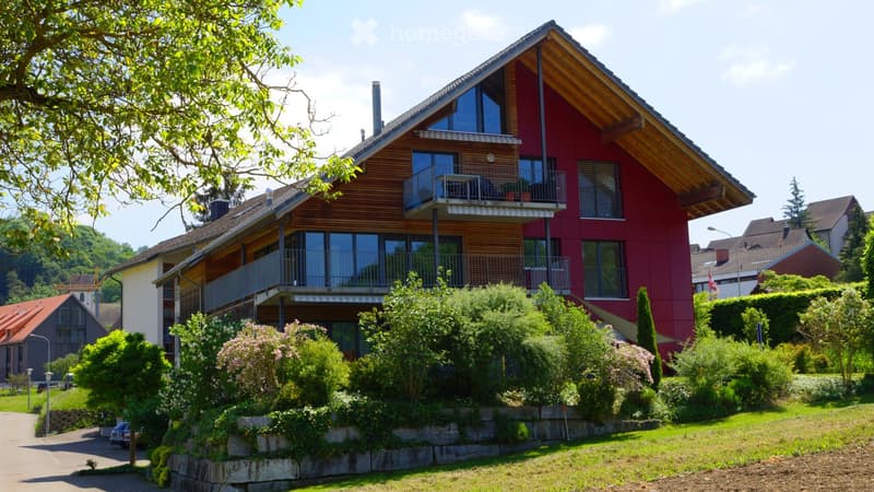 Modern two-story house with red and wooden exterior, multiple balconies, surrounded by greenery and trees.