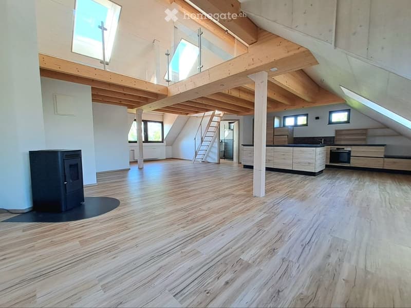 Furnished Room. Open-plan attic space with wooden floors and exposed wooden beams, featuring a modern kitchen area with cabinets and an oven, a staircase leading to an upper level, and a fireplace.