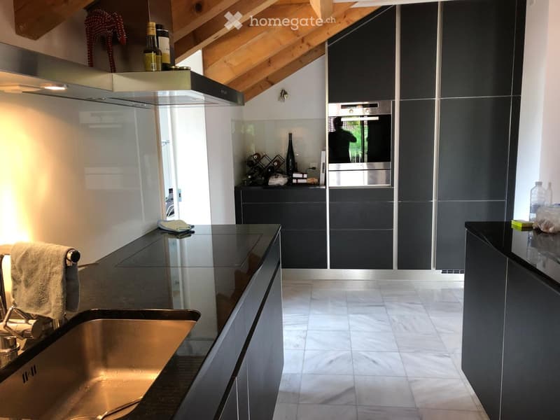 Modern kitchen with dark grey cabinets, stainless steel appliances, and a black countertop. Features include a sink, faucet, oven, wine rack, and decorative bottles.