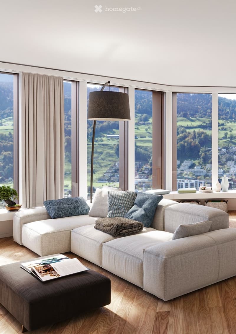 Modern living room with large windows offering a scenic view, a grey sectional sofa with multiple cushions, an ottoman, a floor lamp, a small table with decorative items, and a potted plant.