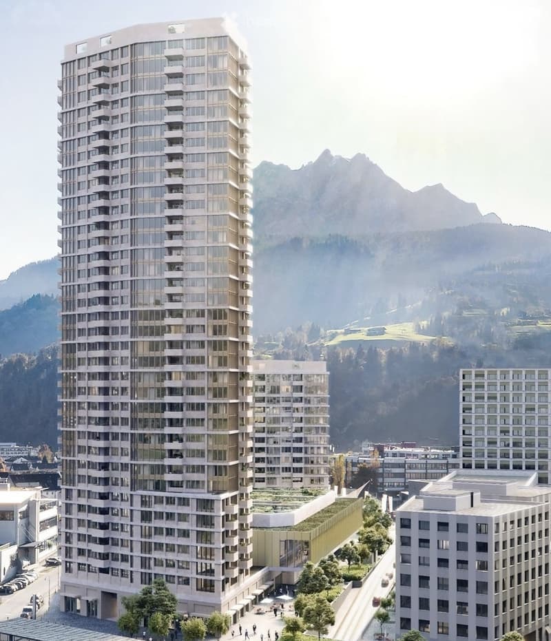 Modern high-rise building with glass and concrete facade, surrounded by smaller structures and green spaces, with mountains in the background.