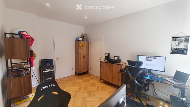 A well-lit room with wooden flooring, a desk setup including a laptop, screen, and chair, a cabinet, a printer, a coffee maker, a guitar amplifier, and a helmet on top of a cabinet.