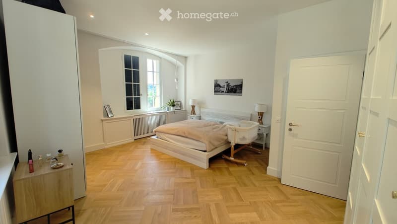 Bright bedroom with wooden flooring, large arched window, bed with beige bedding, nightstand, chair, table lamp, picture frame, and plant on windowsill.
