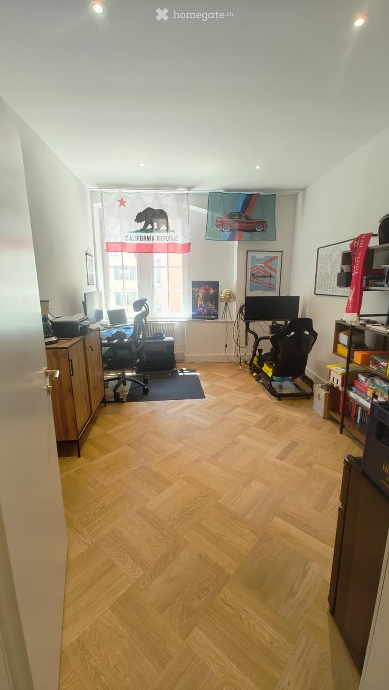 A well-lit room with wooden flooring, a desk setup with a chair, a gaming chair, and shelves with various items. The room features large windows with curtains and artwork on the walls.