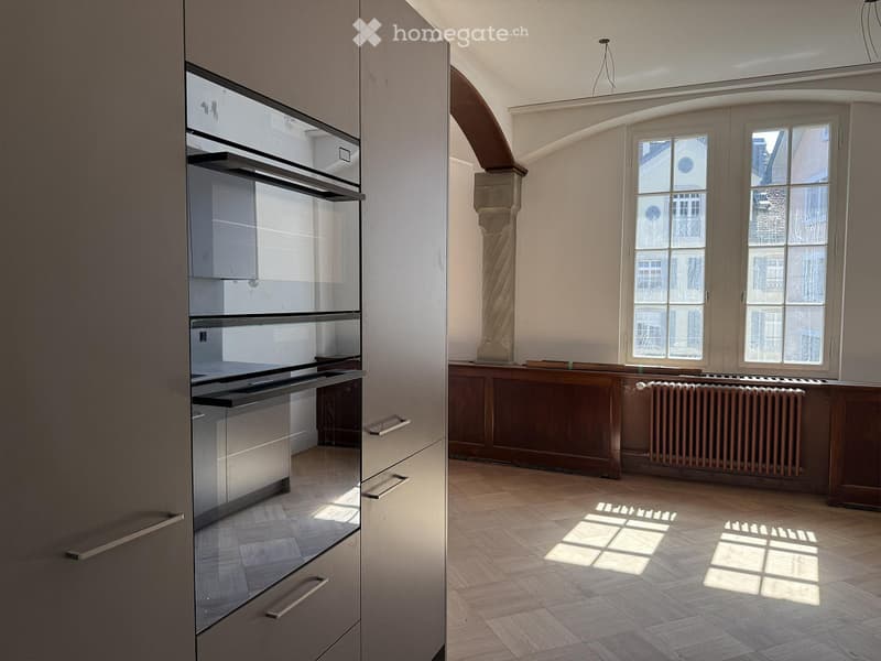 Bright, unfurnished room with wooden flooring, large windows, and a modern kitchen area featuring stainless steel appliances and cabinets.