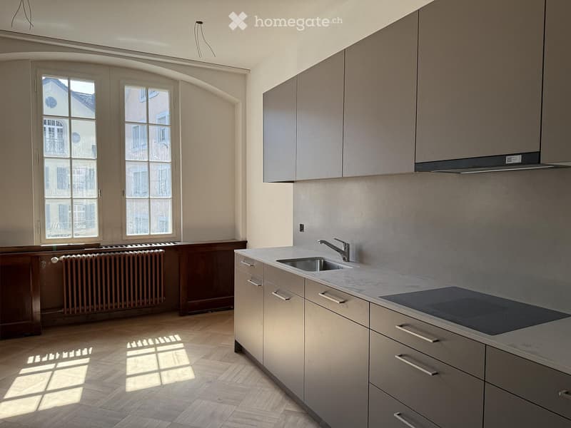 Modern kitchen with light grey cabinets, a stainless steel sink, and a built-in stove. The space features wooden flooring and large windows allowing natural light to fill the room.