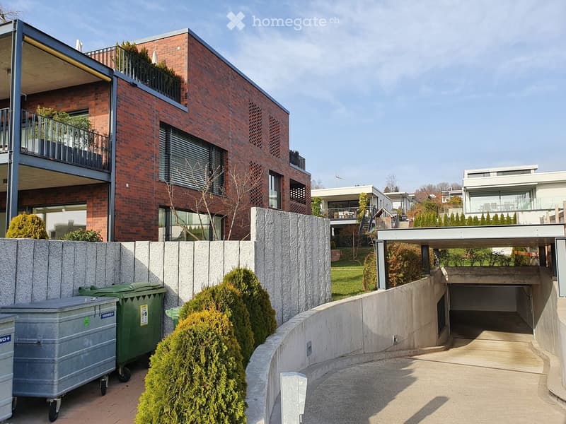 Modern two-story brick building with balconies, surrounded by greenery and a concrete pathway leading to a garage entrance.