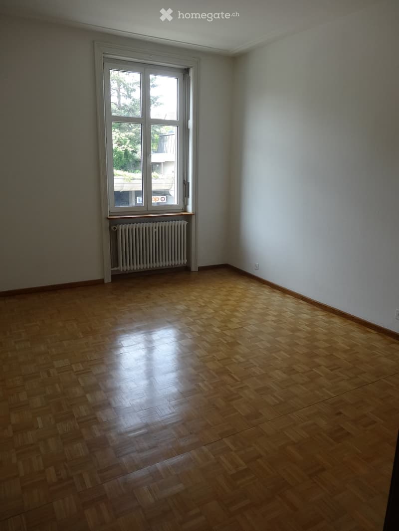 Empty room with wooden parquet flooring, white walls, and a large window with a radiator below it.