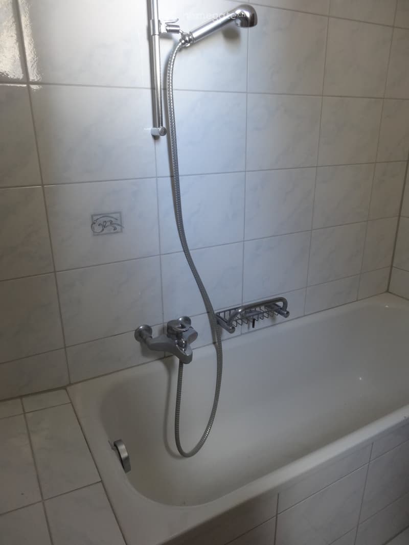 Bathtub with a handheld showerhead and hose, mounted on tiled walls. Includes a built-in shower rack and a standard faucet.