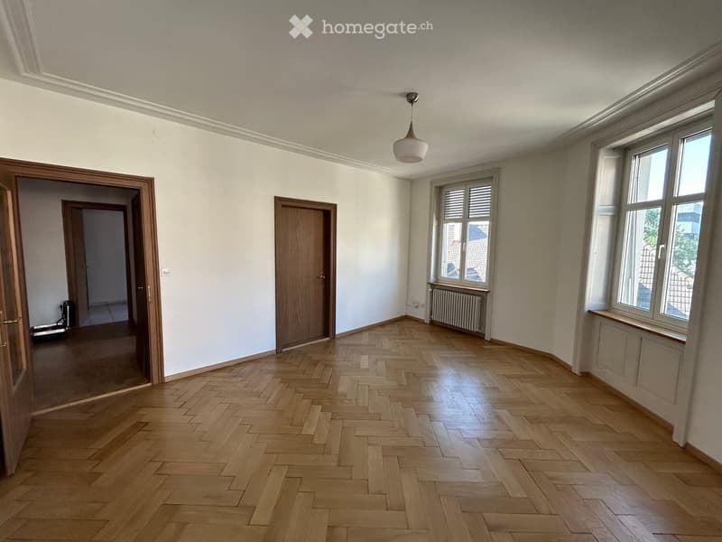 Empty room with wooden herringbone flooring, white walls, large windows, and a hanging light fixture.